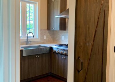 A dark brown barn door, halfway open showing the kitchen with a stainless steel sink and light grey-brown cabinets.