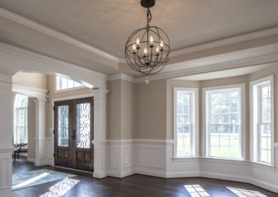 Living room area with light fixture, dark brown floors and front door. A large window brings in natural light.